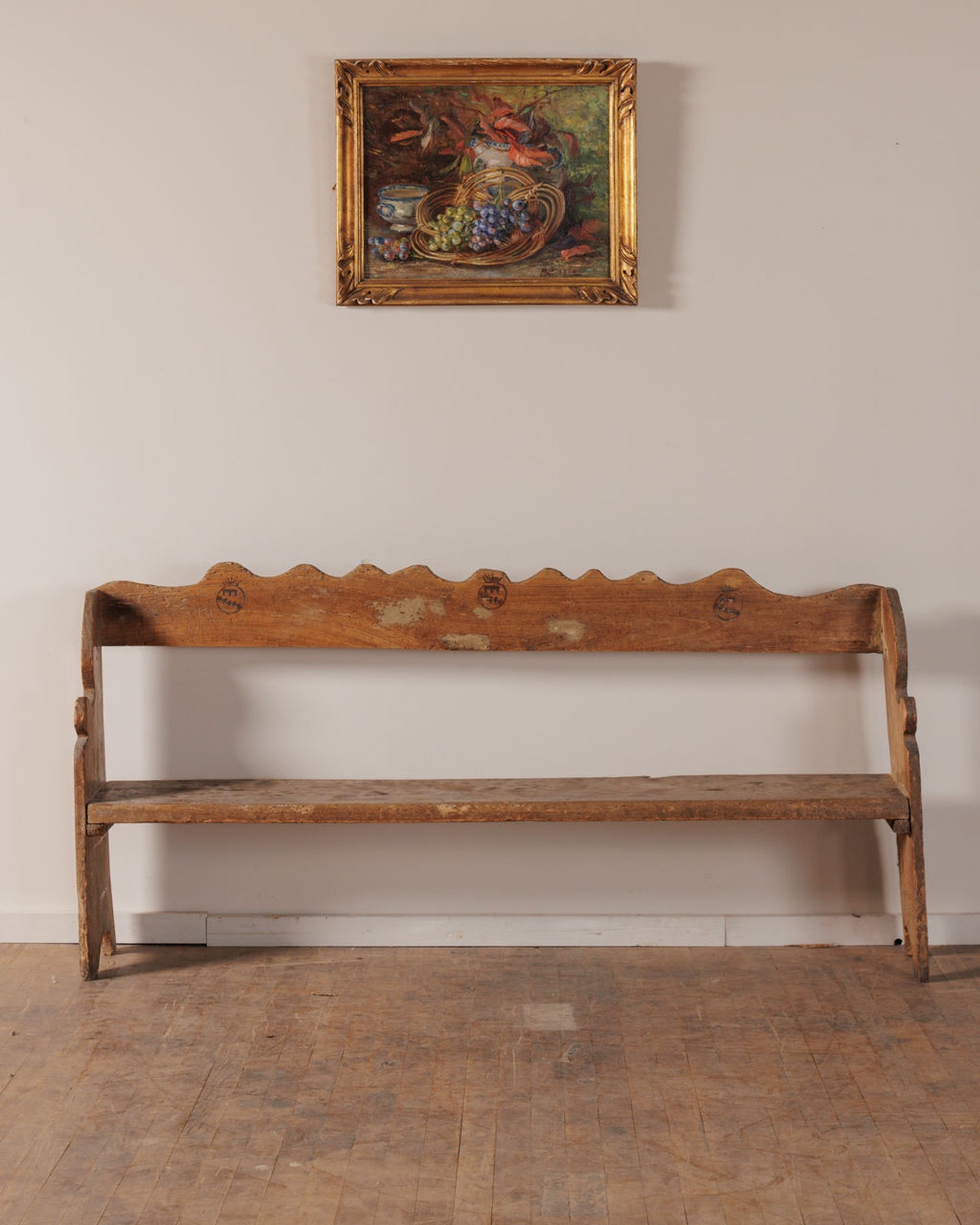 A rustic wooden bench with a scalloped backrest below a framed still life painting of grapes.