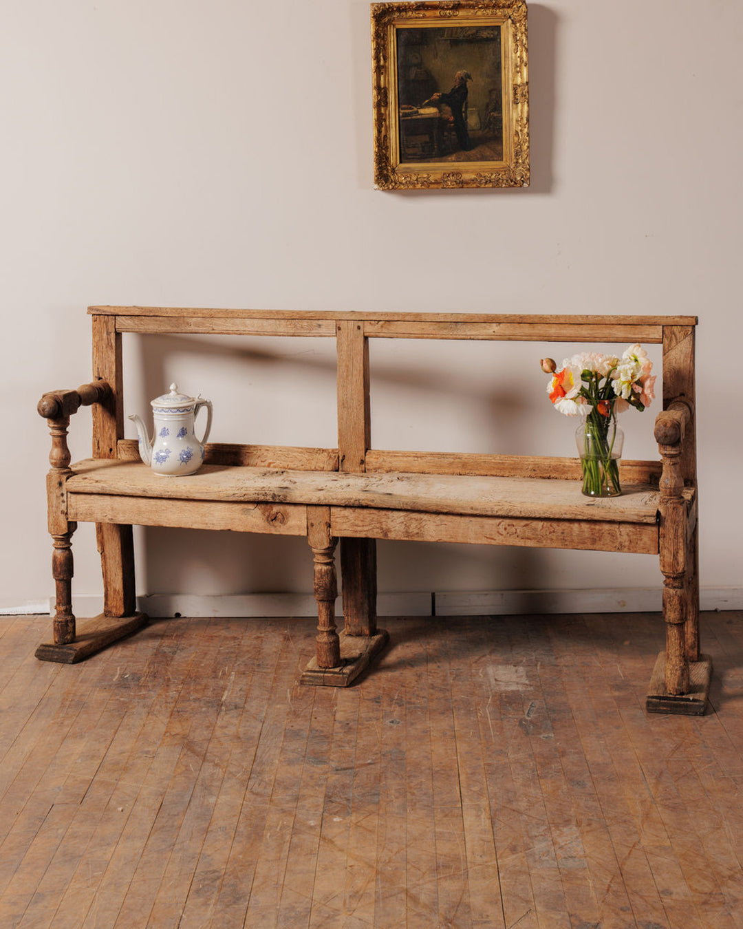 An antique rustic wooden bench with a blue and white teapot and colorful flowers, beneath a framed painting.