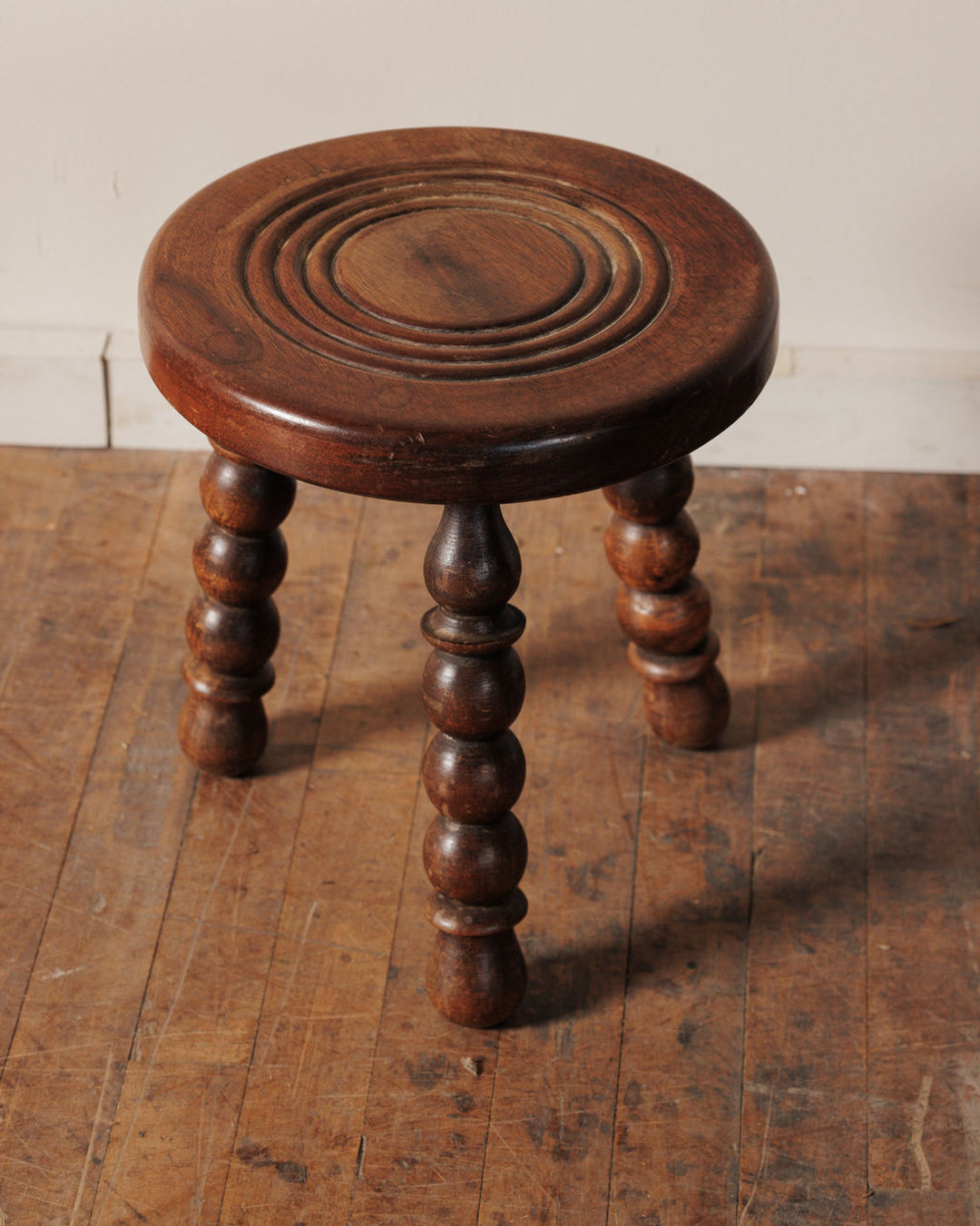 Dark wooden stool with a round seat featuring concentric carved rings and three bobbin-turned legs.