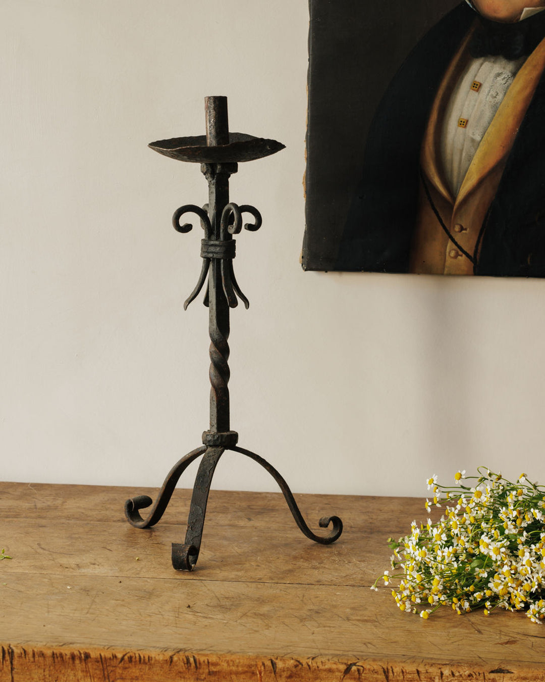 Wrought iron candlestick on a wooden table with chamomile flowers and a partial portrait painting.