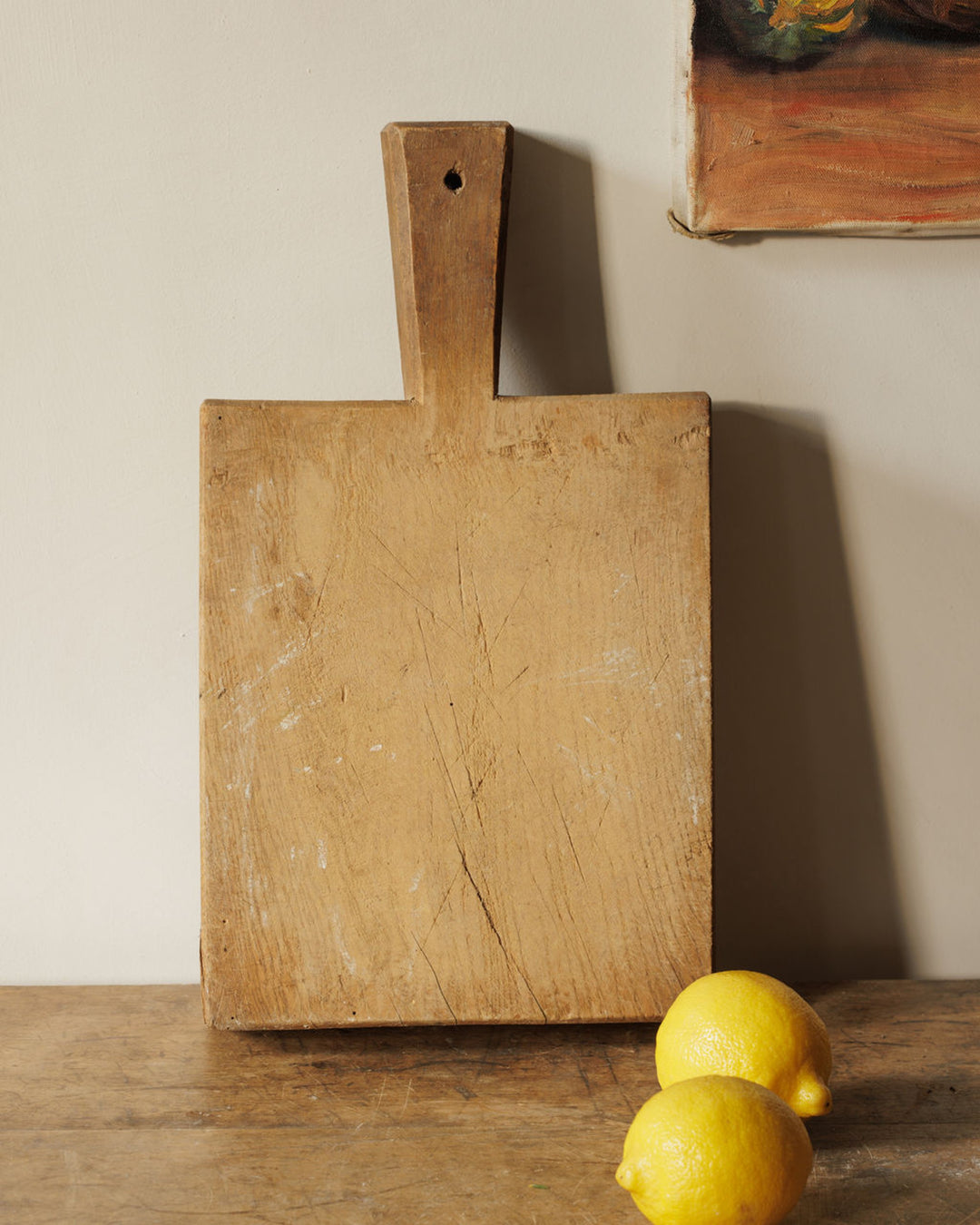 An old wooden cutting board with a handle stands upright, with two lemons on a wooden table.