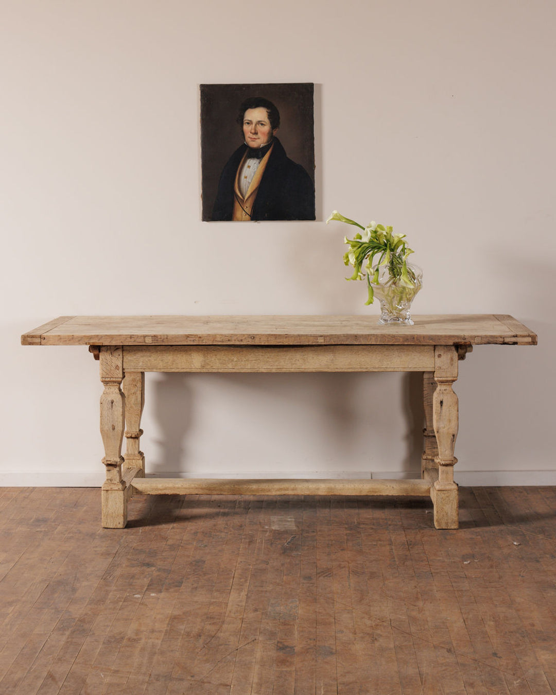 A rustic wooden table with a vase of white flowers, beneath a portrait of a man in a dark coat and yellow vest.