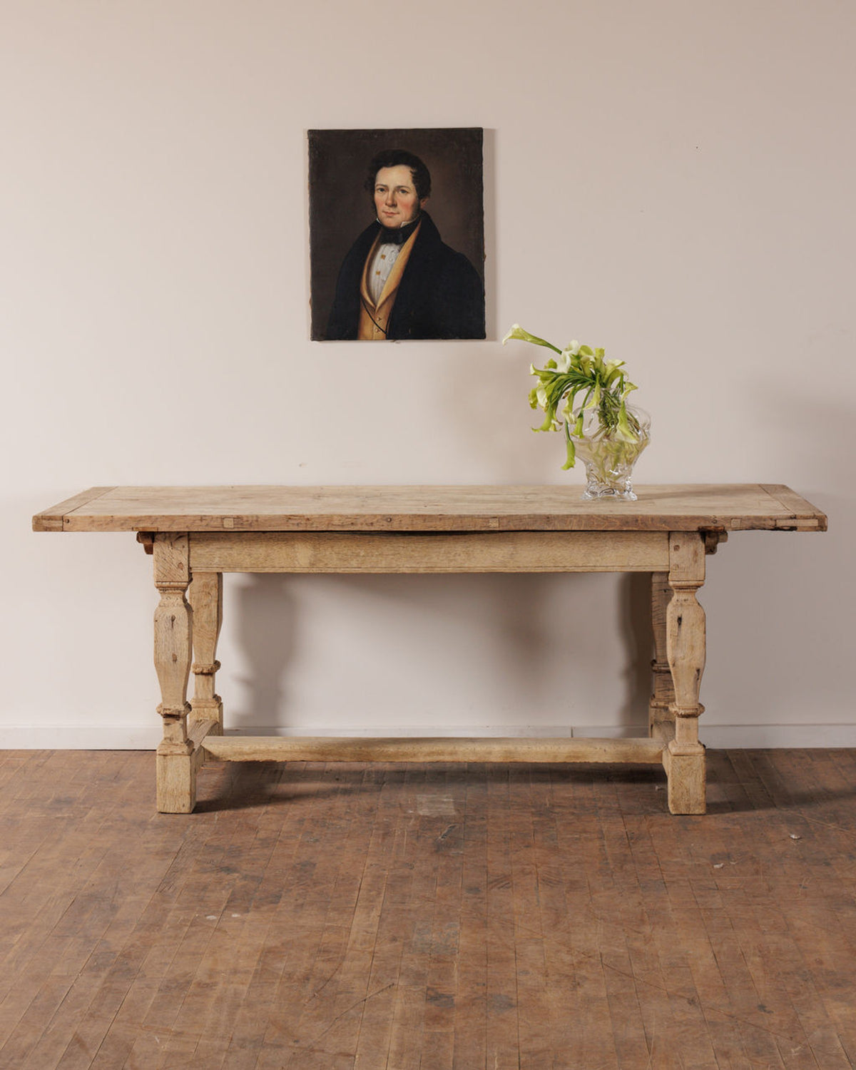 A rustic wooden table with a vase of white flowers, beneath a portrait of a man in a dark coat and yellow vest.