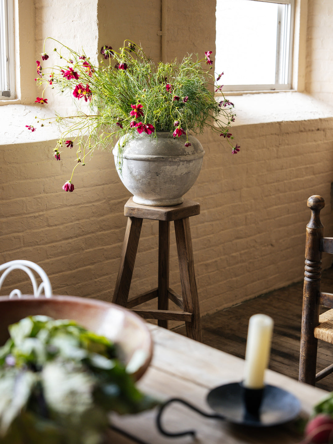 a Willy Guhl pot filled flowers sits on a wooden stool in front of a window .
