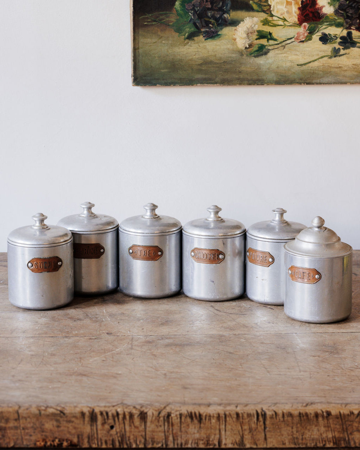 Six vintage silver-toned kitchen canisters labeled for items like Sugar, Flour, Tea, and Coffee, on a wooden table.