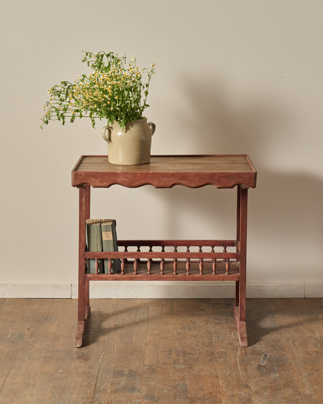A rustic red-toned wooden side table with a vase of white and yellow wildflowers on top and books on a lower spindle shelf.
