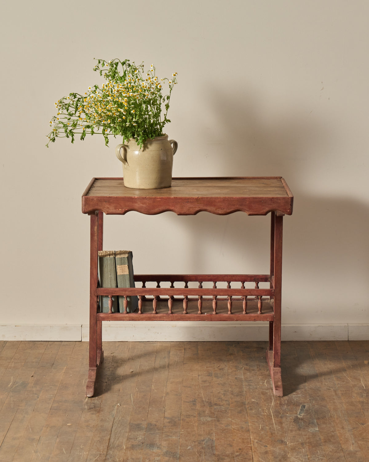 A rustic red-toned wooden side table with a vase of white and yellow wildflowers on top and books on a lower spindle shelf.