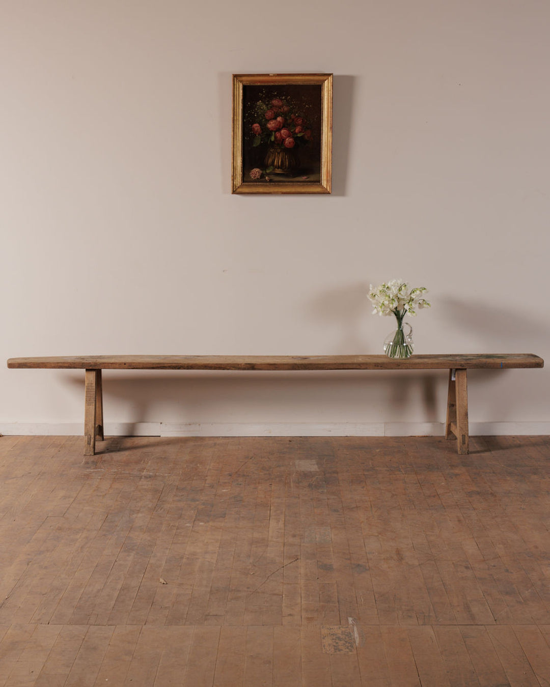 A long rustic wooden bench with a vase of white flowers, below a framed floral still life painting on a light wall.
