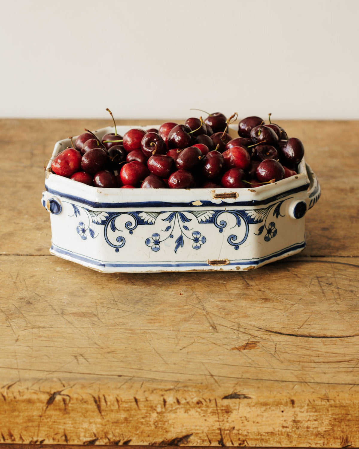 White octagonal bowl with blue floral patterns filled with dark red cherries on a wooden table.