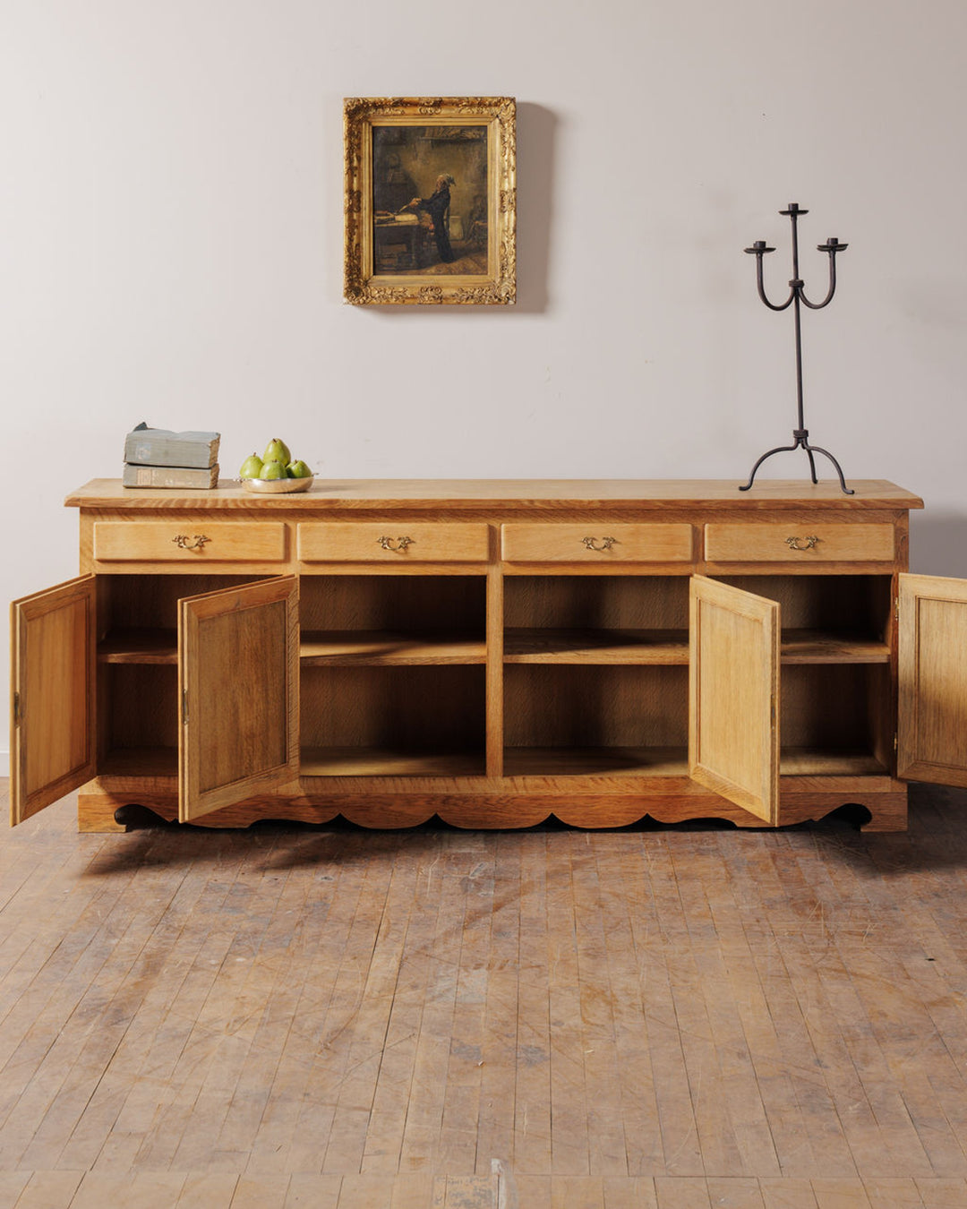 A long, light wooden sideboard with four drawers and open cabinet doors revealing shelves. On top are pears, stacked gray blocks, and a black candelabra, with a framed painting hanging above on a light wall.