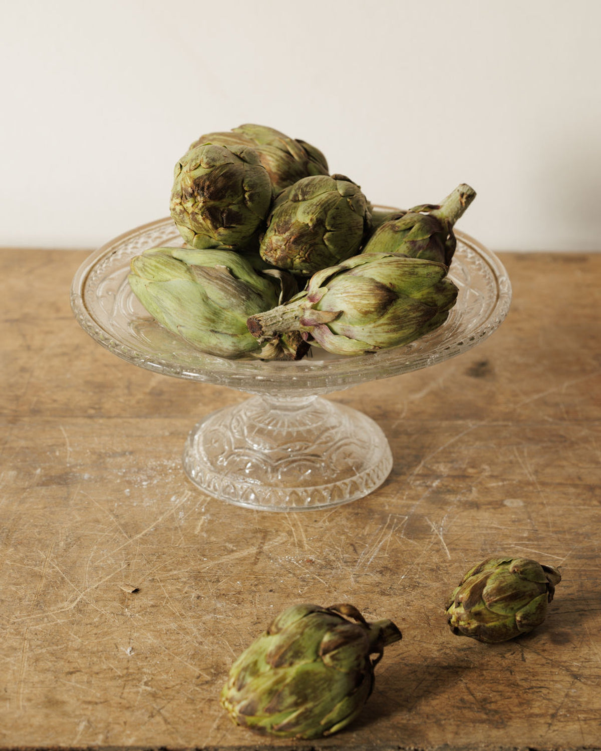 Green and brown artichokes on a glass cake stand and a wooden table.