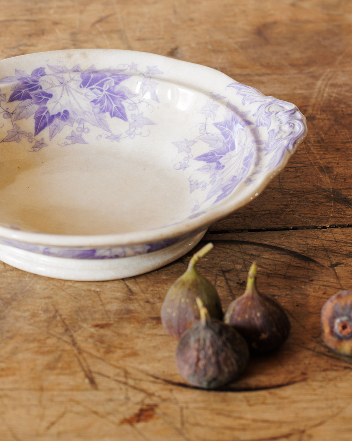 A white bowl with purple ivy leaf patterns on a wooden surface next to several figs.