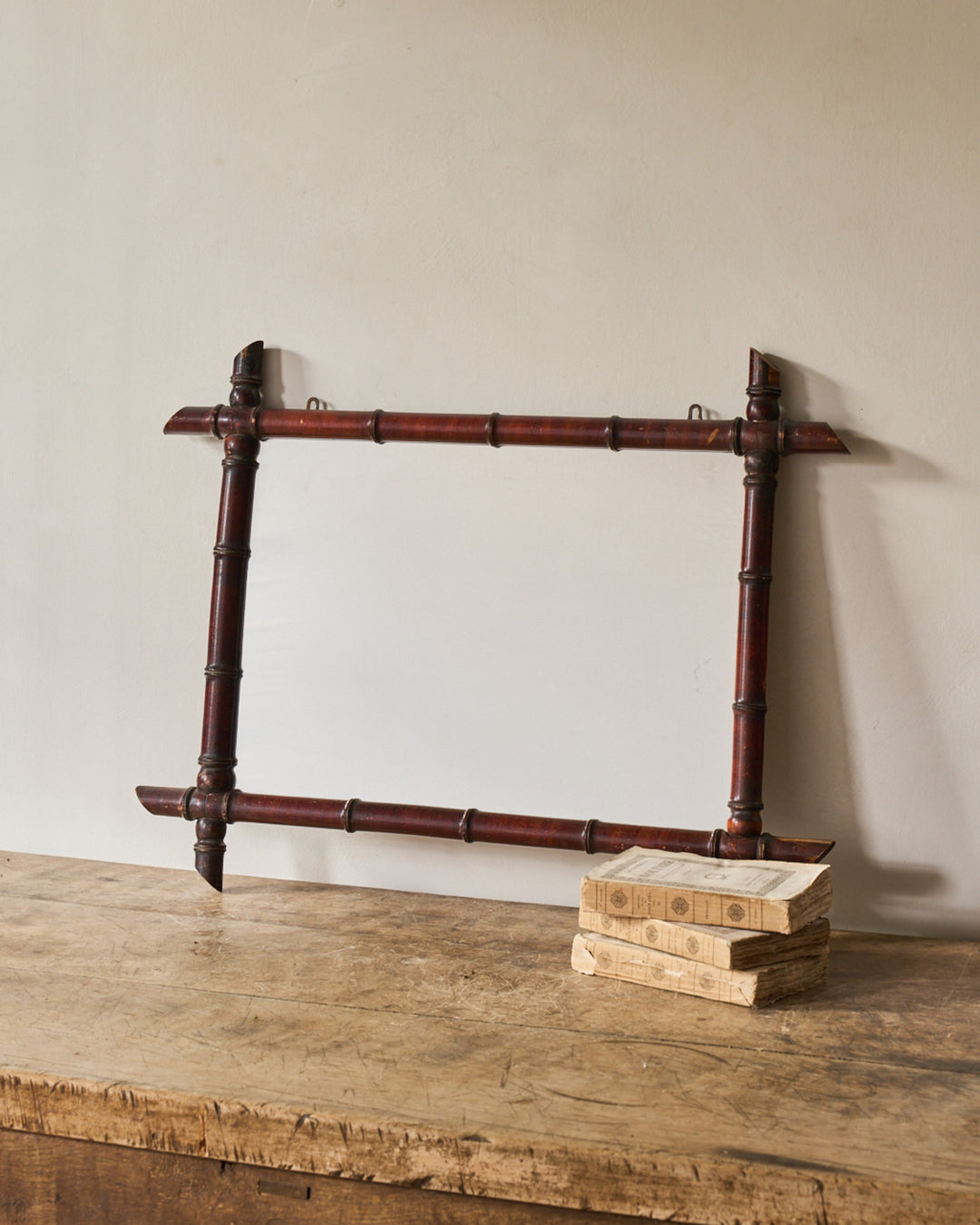 A rectangular, dark red faux bamboo mirror frame rests on a distressed wooden table next to a stack of old books.