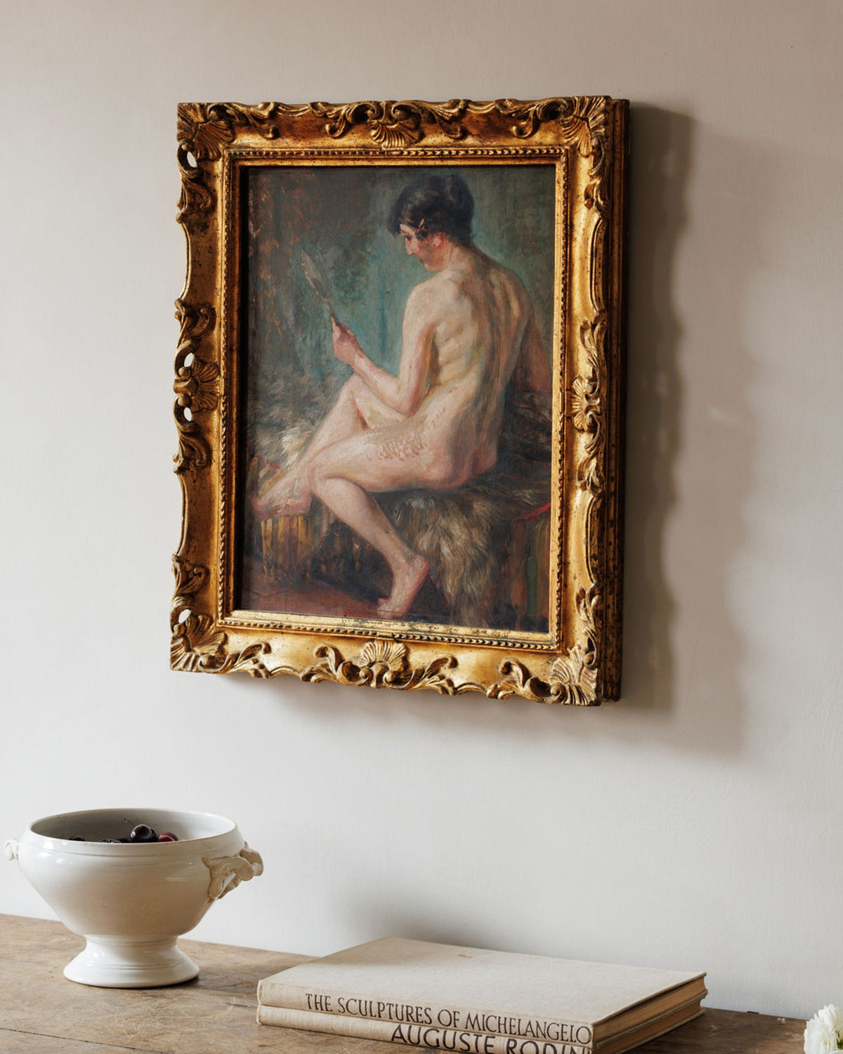 An ornate gold-framed painting of a seated nude woman looking into a handheld mirror hangs on a light wall, with a white bowl of berries and books on a table below.