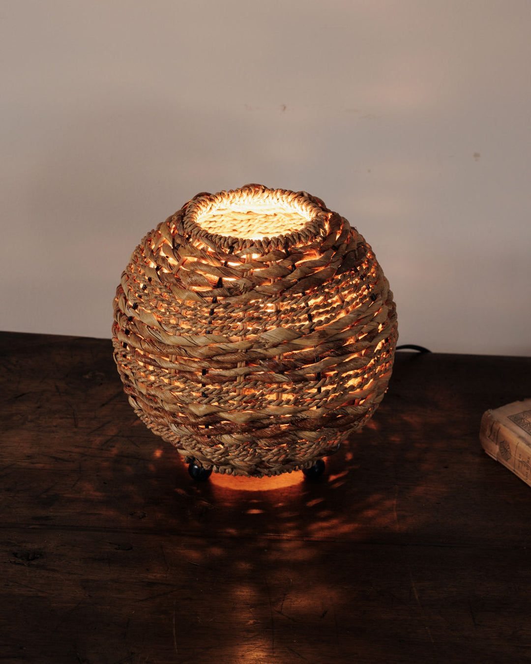 Round woven straw lamp glowing on a dark wooden table.