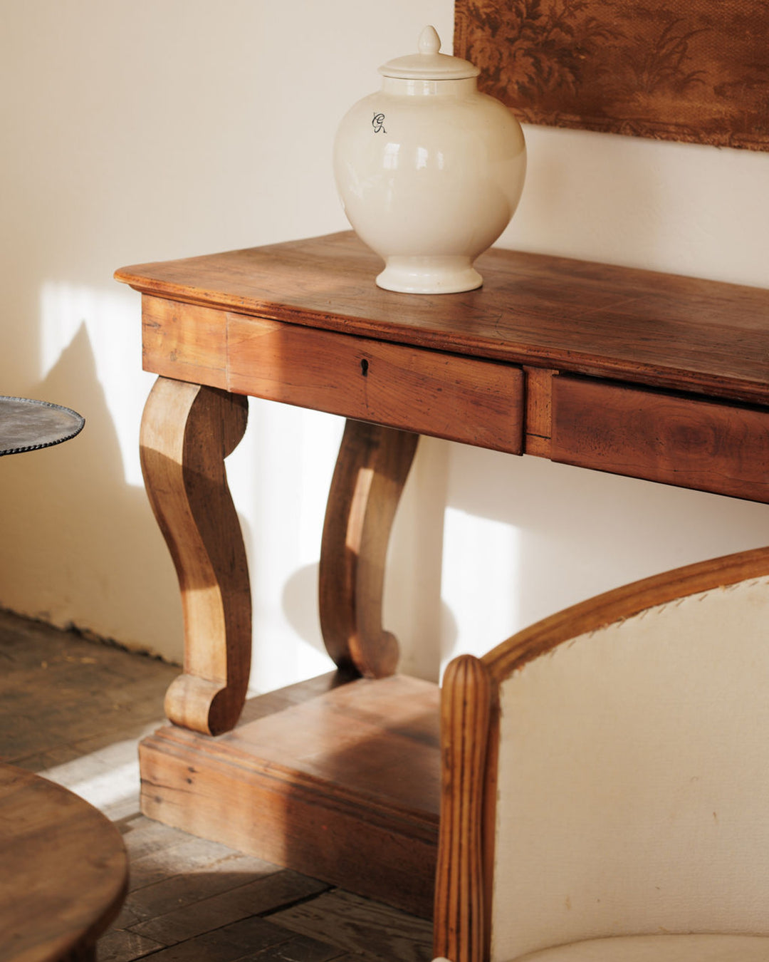 A vintage white lidded jar sits on a dark wood console table, lit by sunlight, with an antique upholstered chair in the foreground.