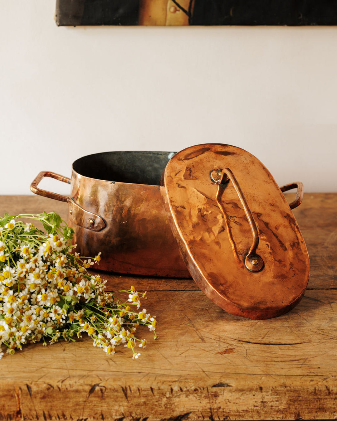 Vintage oval copper pot with lid and wildflowers on a rustic wooden table.