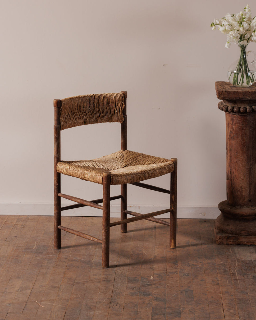 A rustic wooden chair with a rush seat and back, beside a wooden pedestal with a vase of white flowers.