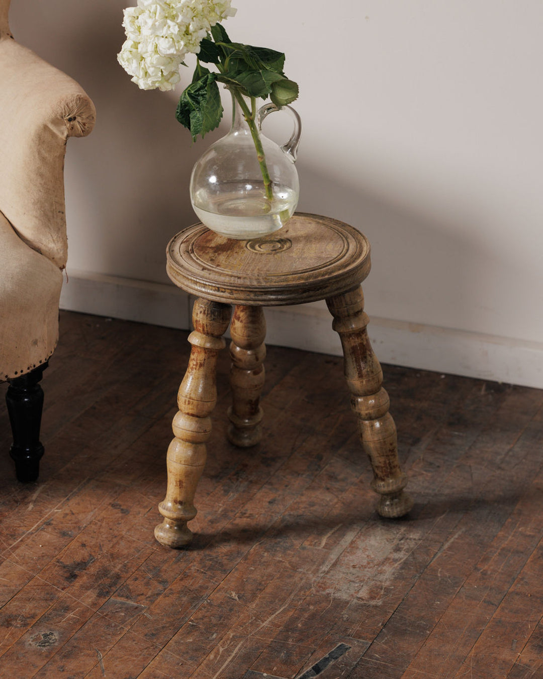 Vintage wooden stool with a vase of white flowers next to an armchair on a rustic wood floor.