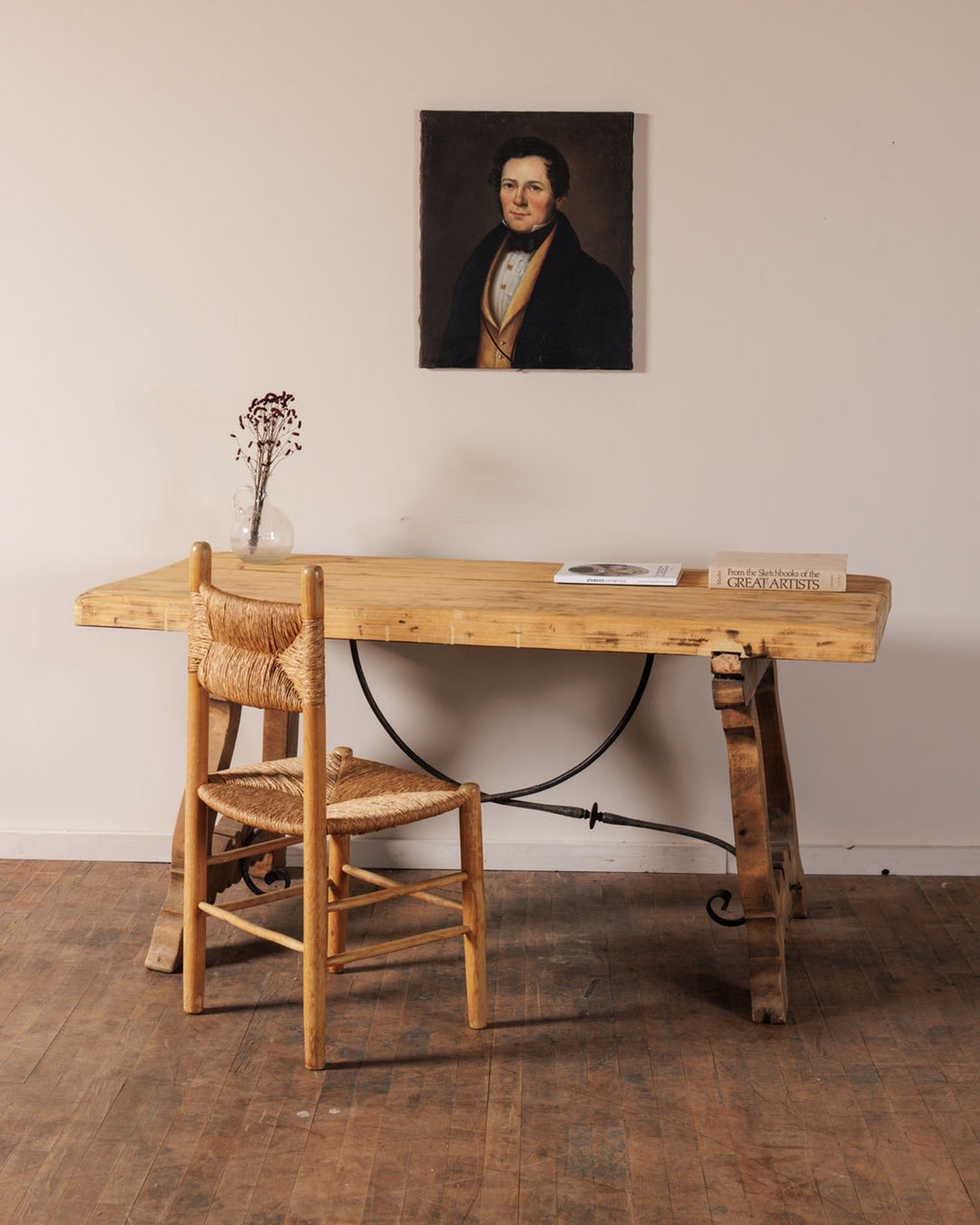 A rustic wooden desk with a rush-seated chair, a vase of dried flowers, and books. A portrait of a man hangs on the light wall above.