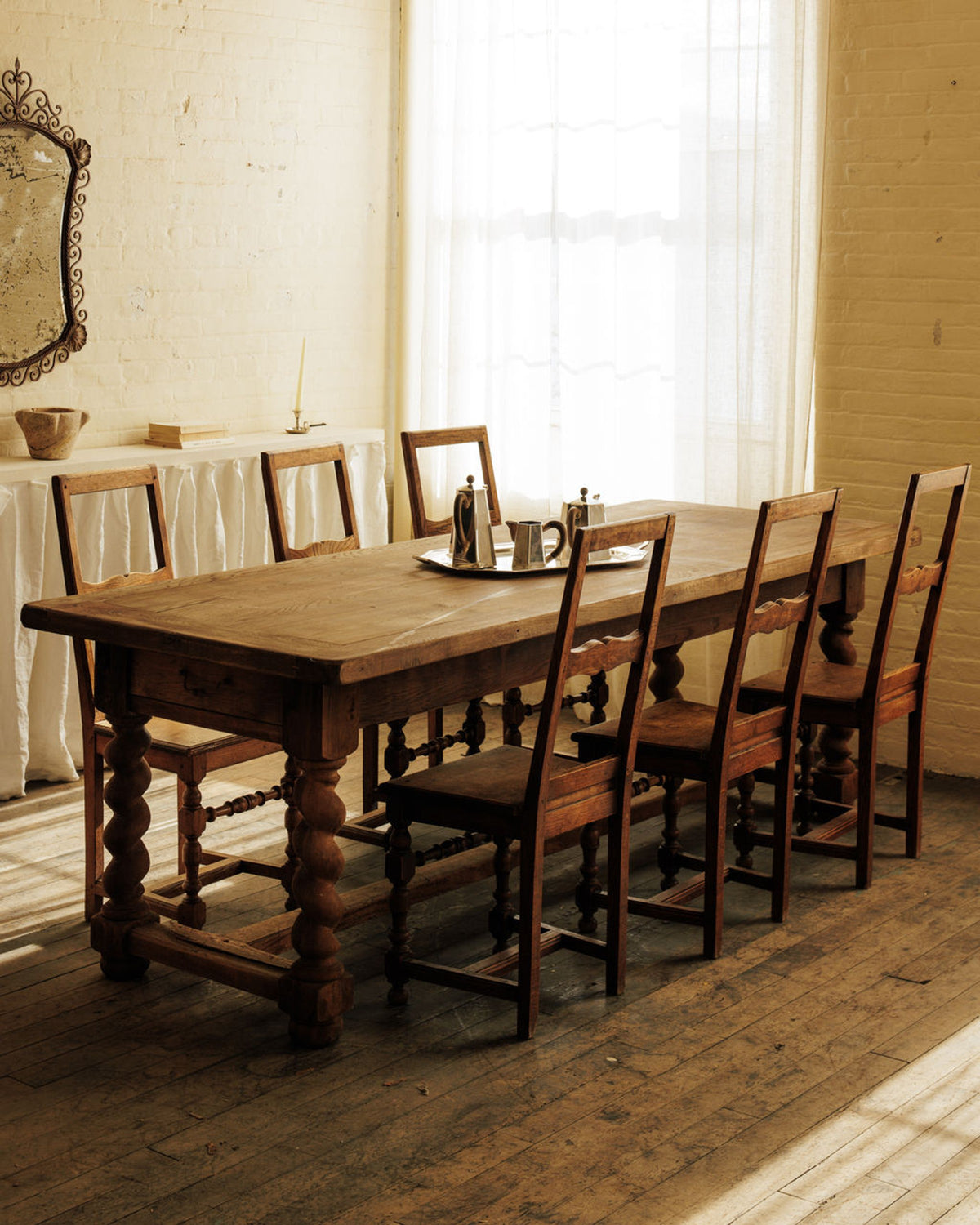 Rustic room with an antique wooden table, six chairs, and silver pitchers on a tray.