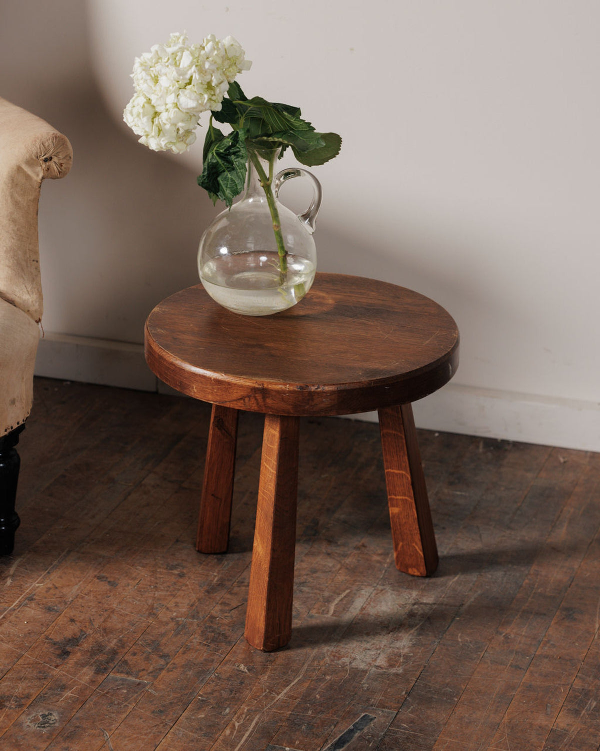 A round wooden stool with a vase of white hydrangeas on a wooden floor.