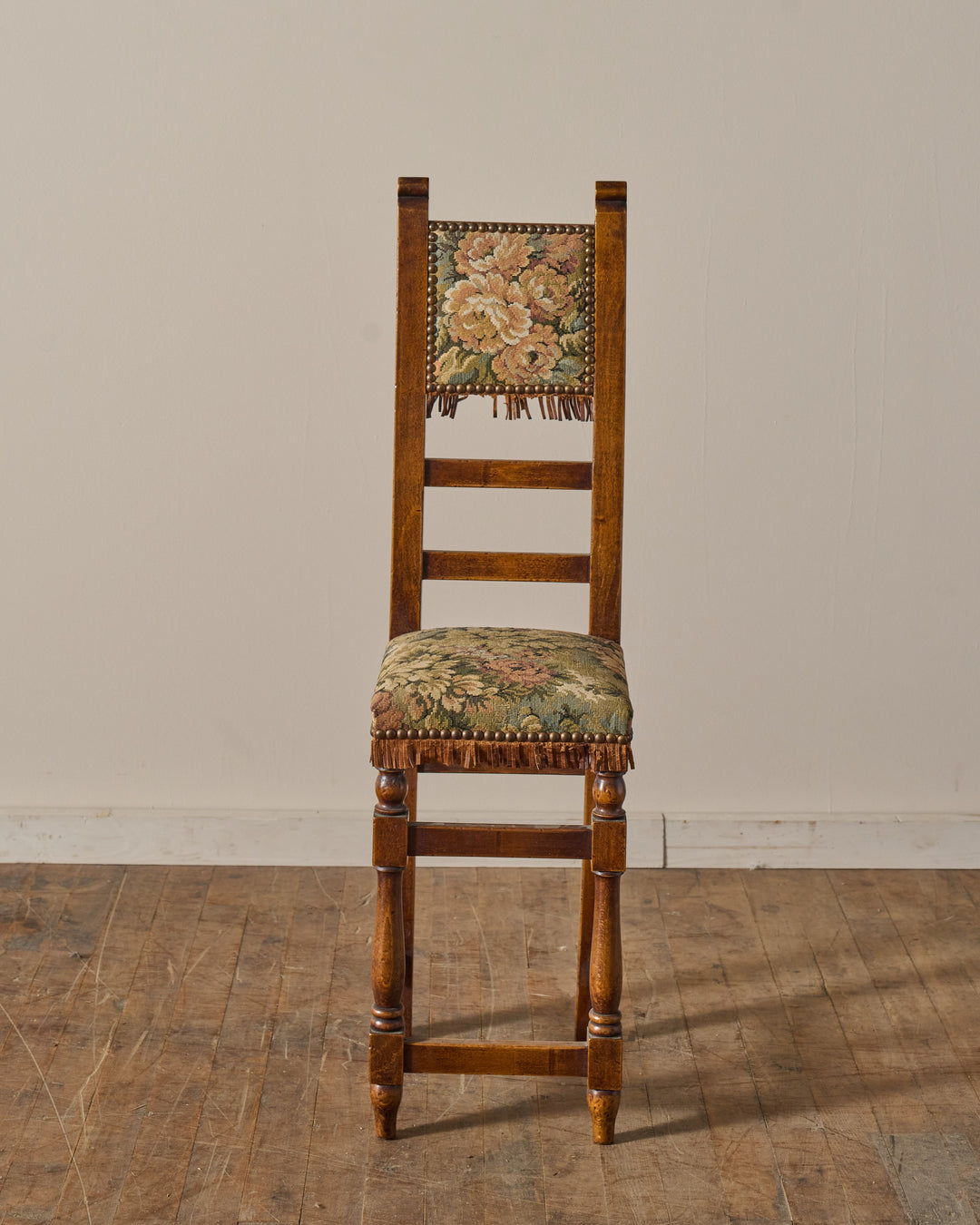 Tall wooden chair with floral tapestry upholstery, fringe trim, and nailhead accents.