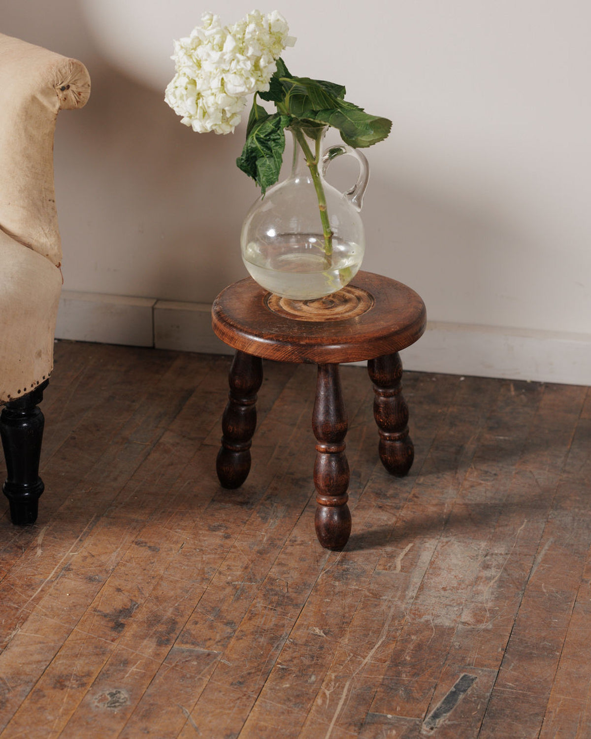 White hydrangeas in a glass pitcher on a dark wooden stool, next to an upholstered chair on a distressed wood floor.