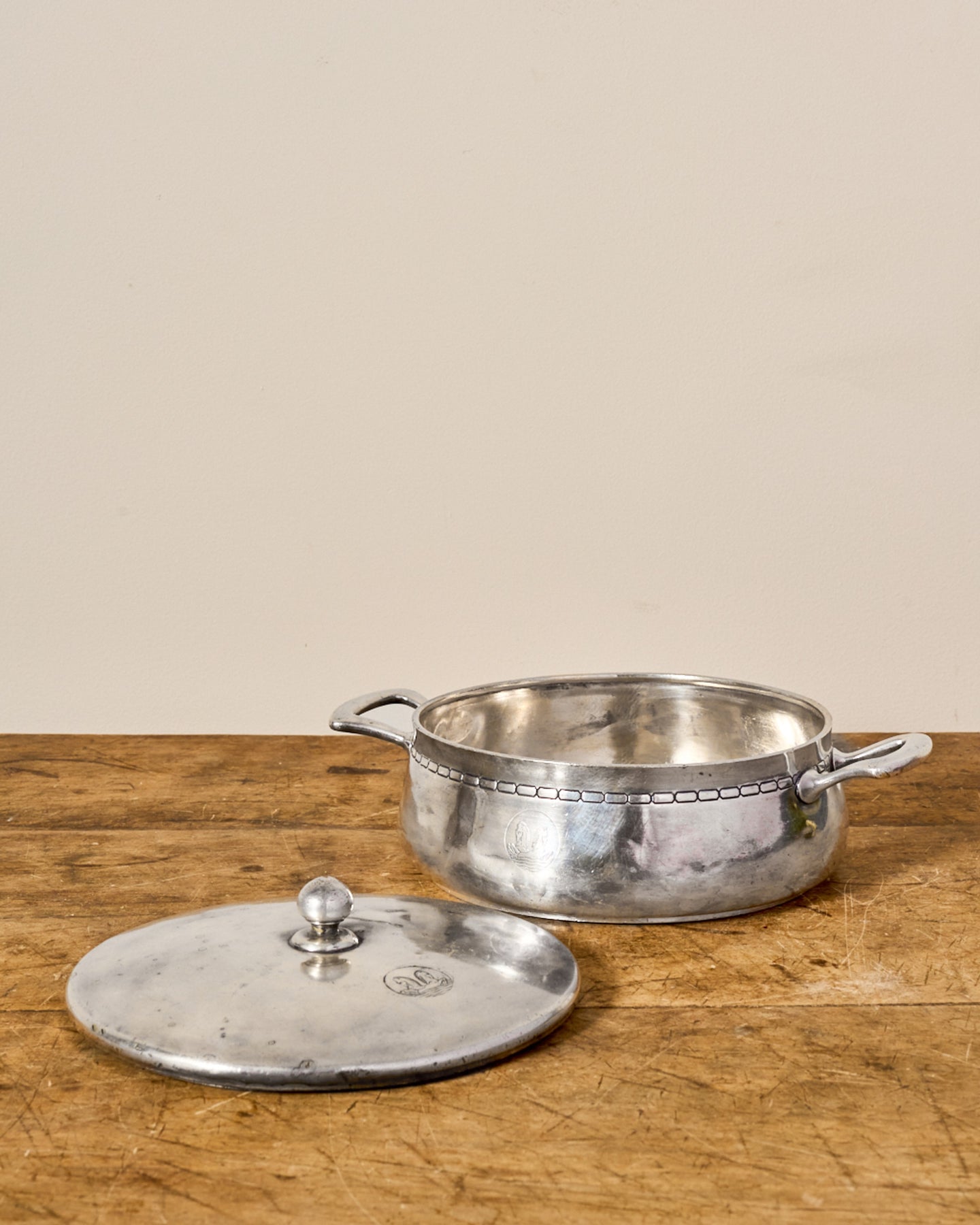 A vintage metallic pot with handles, with its lid resting beside it on a wooden table.