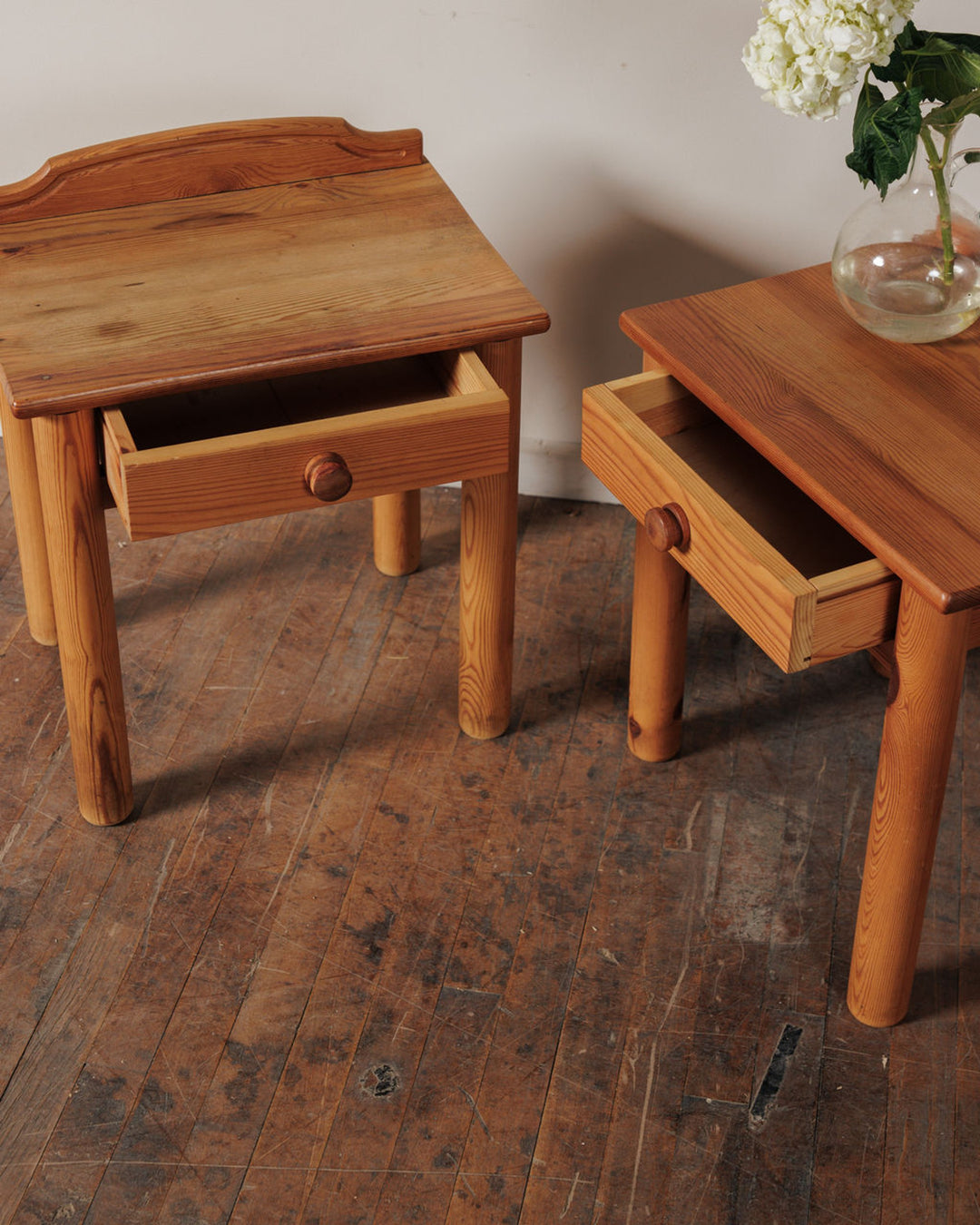Two light wood end tables with open drawers and rounded legs on a wooden floor. A vase of white flowers sits on the right table.