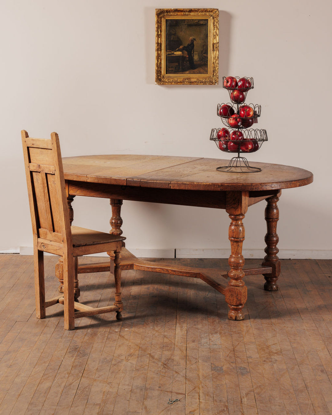 A rustic oval wooden table with a chair, a tiered basket of red apples, and a framed painting on the wall.