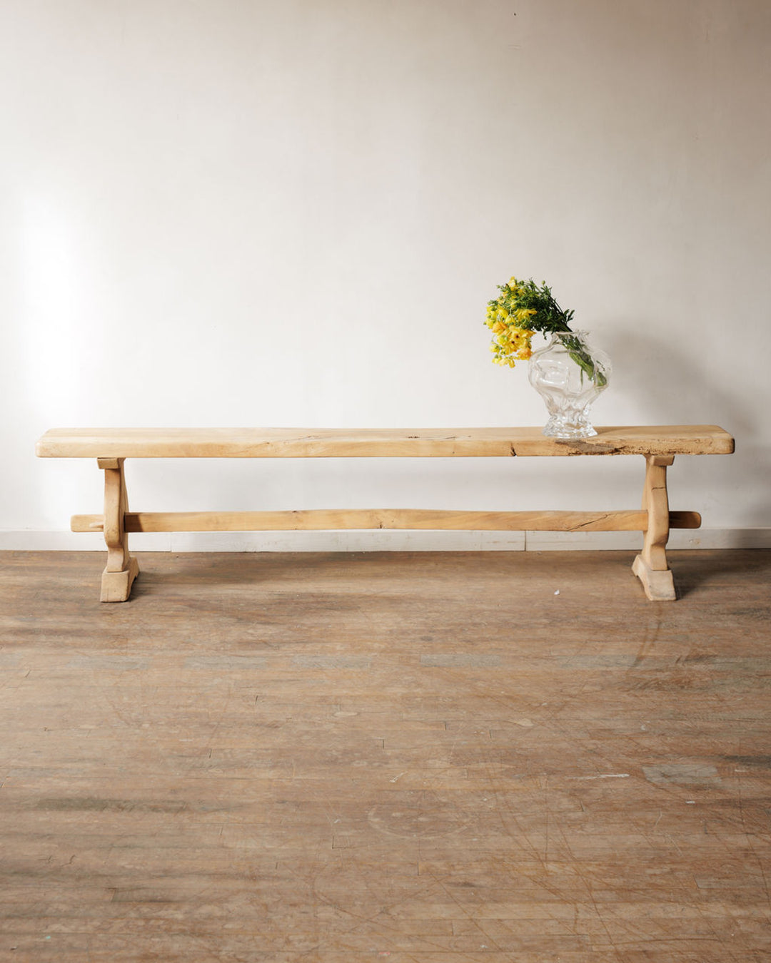 A long, rustic wooden bench with a clear vase of yellow flowers on its right end, against a light wall and wooden floor.