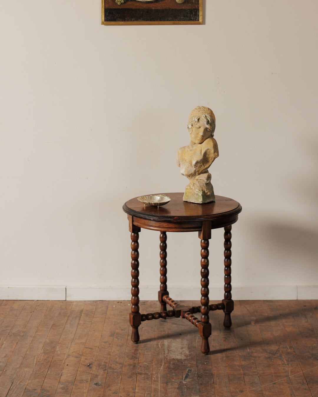 Light-colored bust of a woman and a small bowl on a dark round wooden table with turned legs. A painting is visible above.