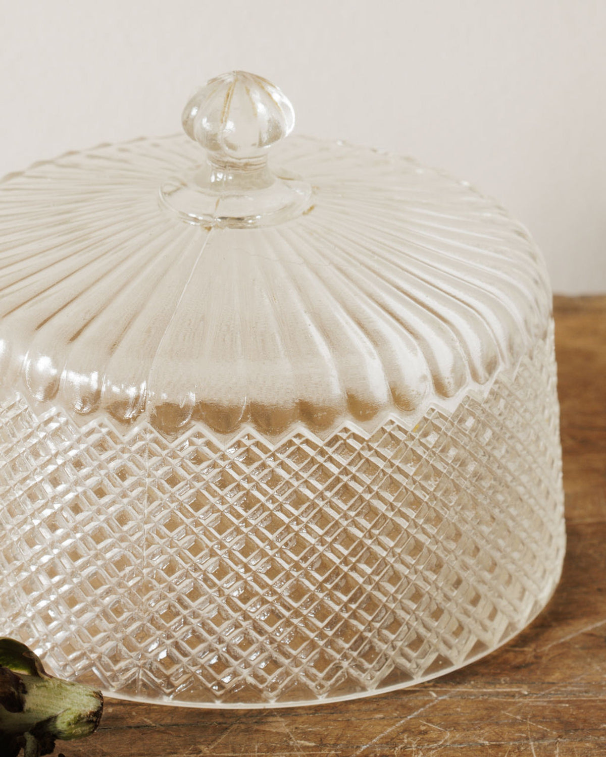 A clear glass cloche with a fluted top and diamond pattern on its sides, sitting on a wooden surface.