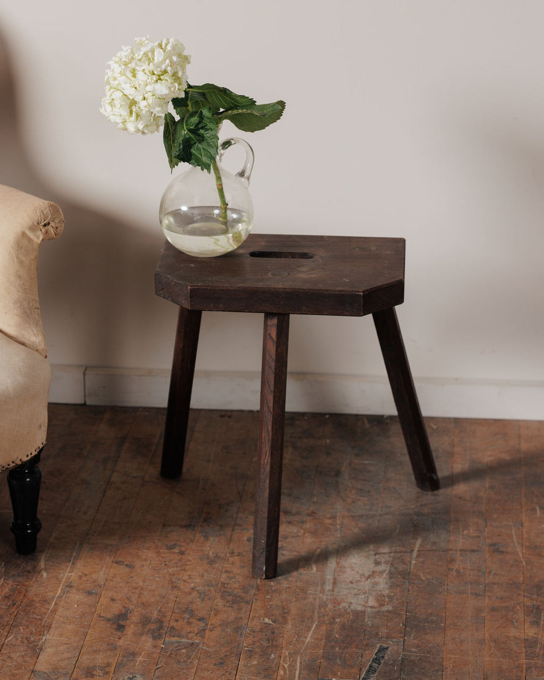 White hydrangeas in a glass pitcher on a dark wooden hexagonal stool.