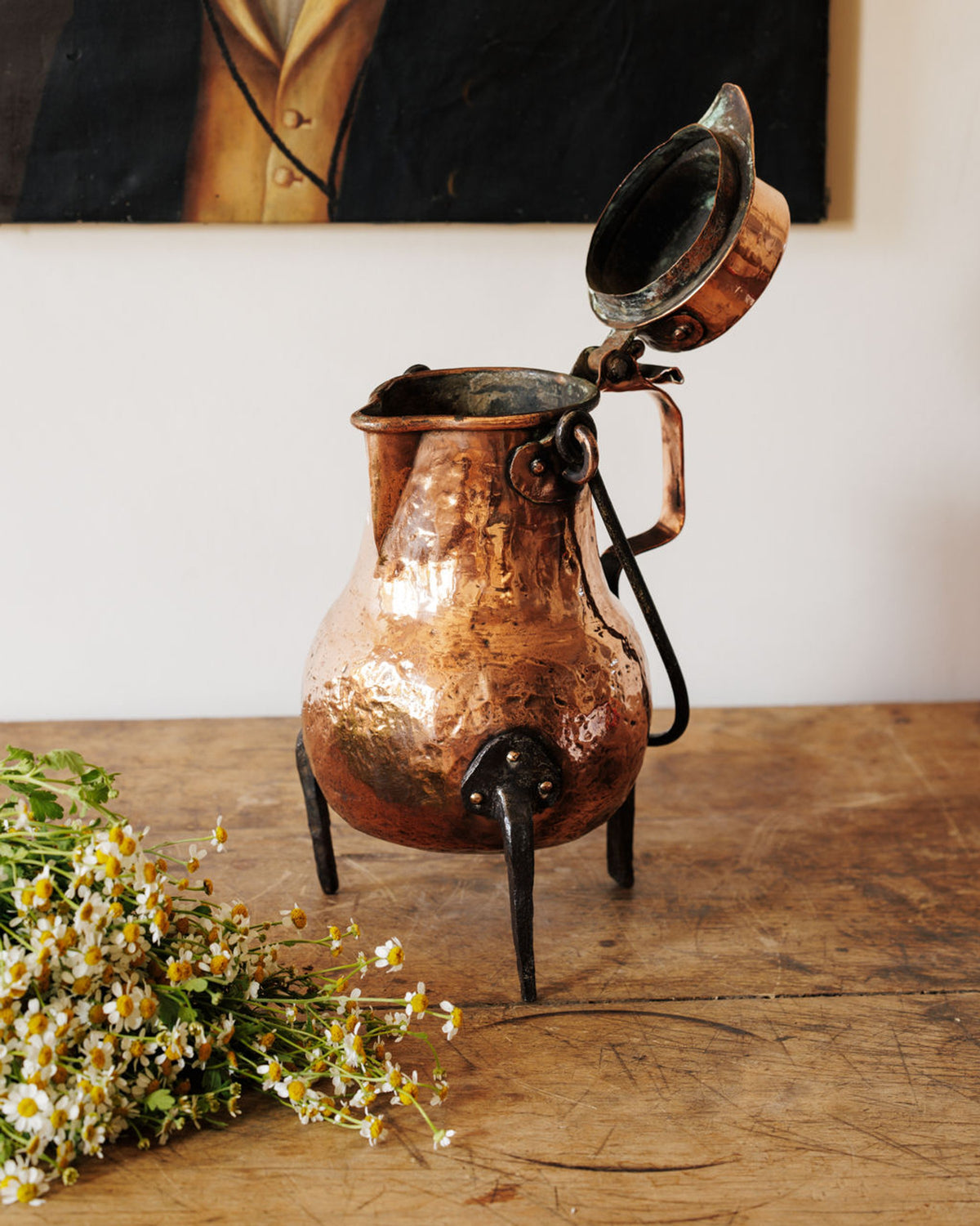 A vintage hammered copper pot with an open lid and three legs sits on a rustic wooden table, next to white wildflowers, with a blurry painting behind.