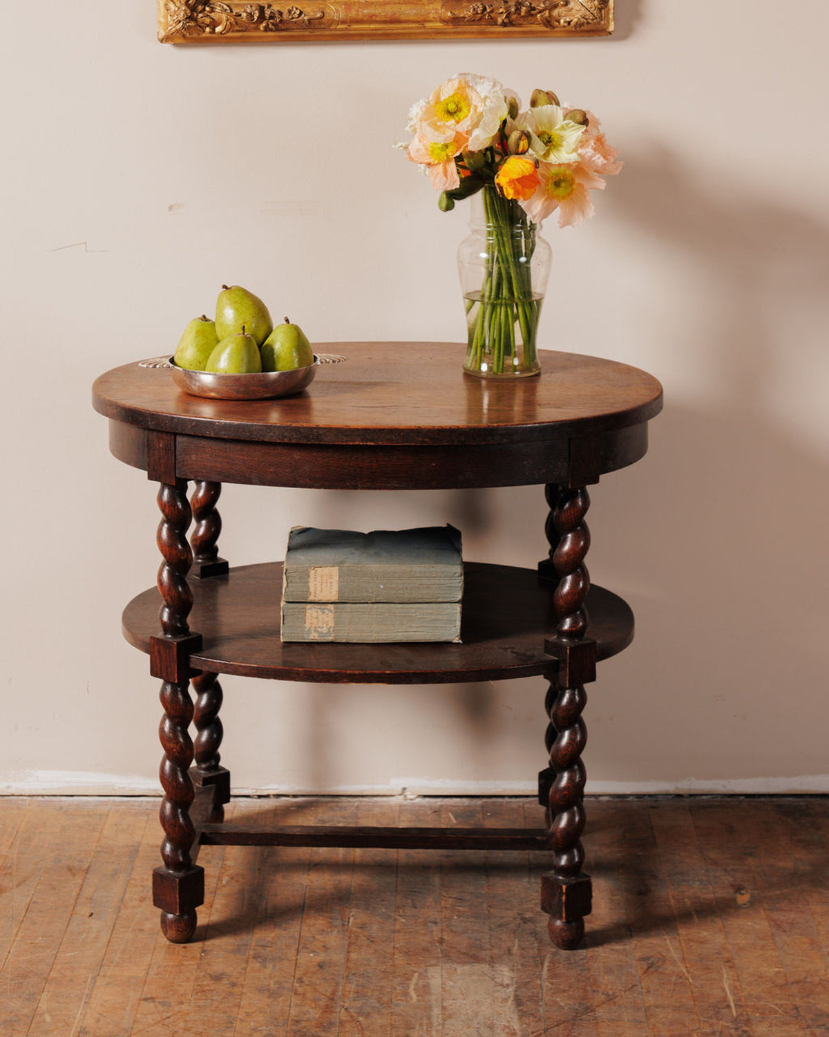 A dark wood, three-tiered round table with barley-twist legs. On top is a silver bowl of green pears and a vase of light orange and yellow flowers. Two old books rest on the middle shelf.