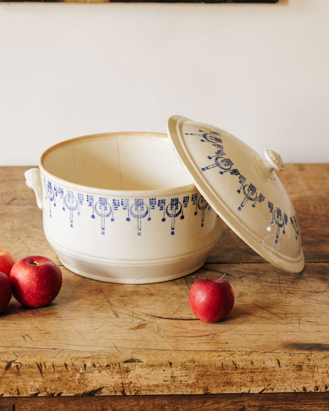 A cream-colored ceramic baking dish with blue patterns, its lid leaning against it, sits on a rustic wooden table with red apples.