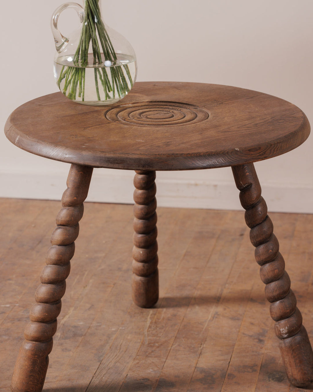 A round wooden stool with twisted legs and a carved swirl on its top, holding a glass pitcher with green stems.