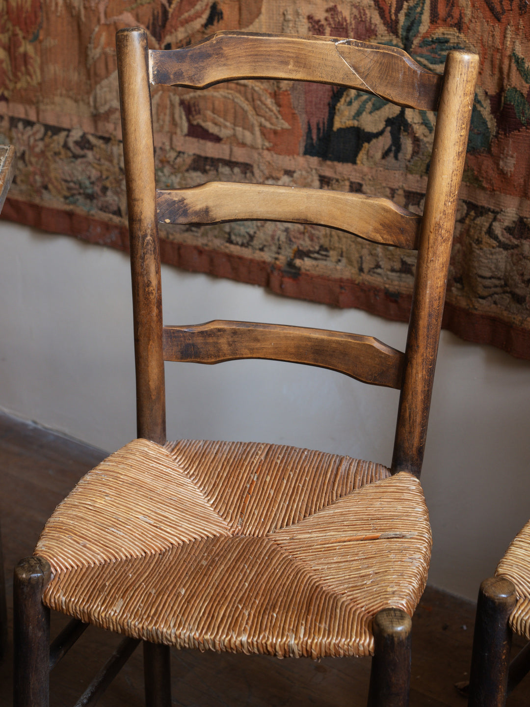 a wooden chair with a wicker seat sits in front of a tapestry