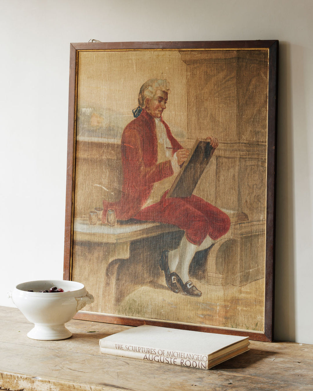 A framed painting of a man in a red coat drawing leans against a wall, with a white bowl of berries and a book titled "The Sculptures of Michelangelo Auguste Rodin" on a wooden surface below.