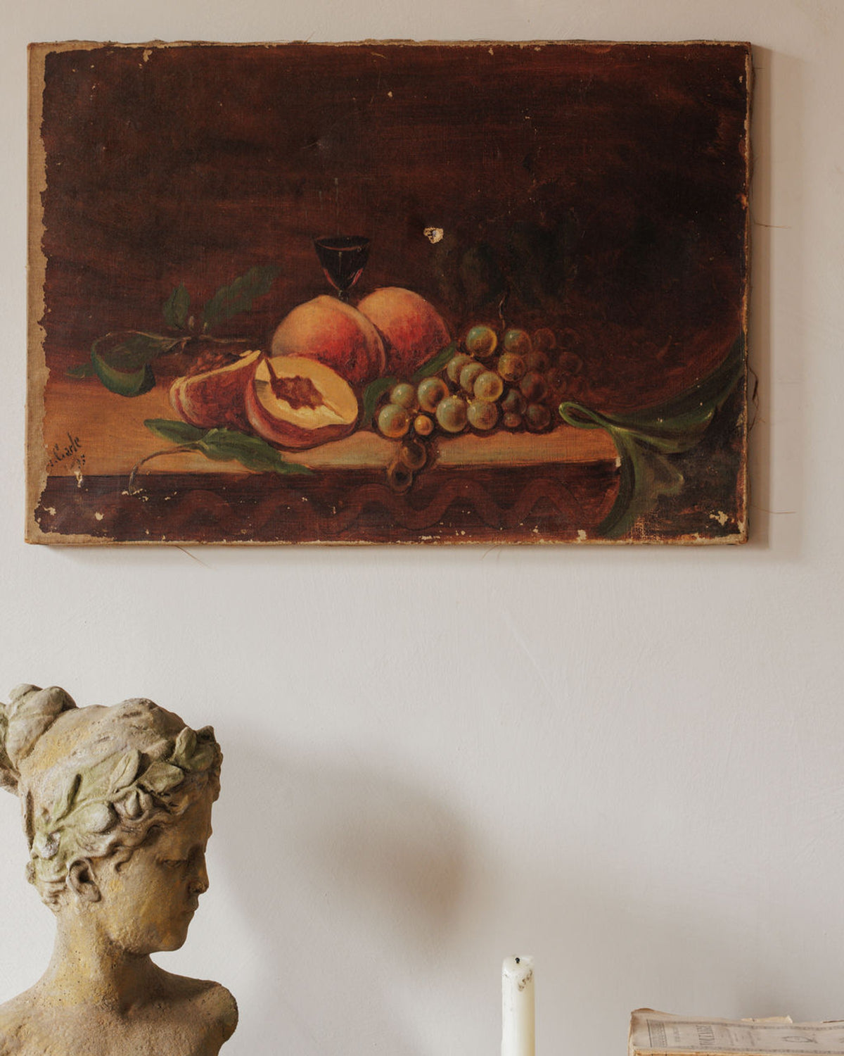 An old still life painting of peaches, grapes, and a wine glass hangs above a classical bust and a candle.