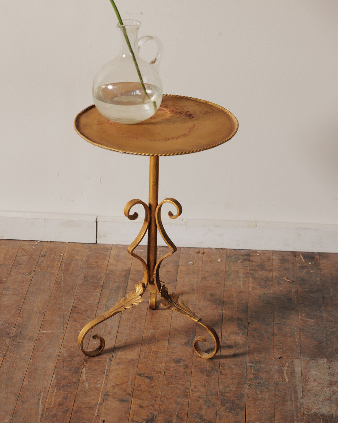 Ornate gold metal side table with a round top and scrolled legs, holding a glass pitcher with a single green stem.