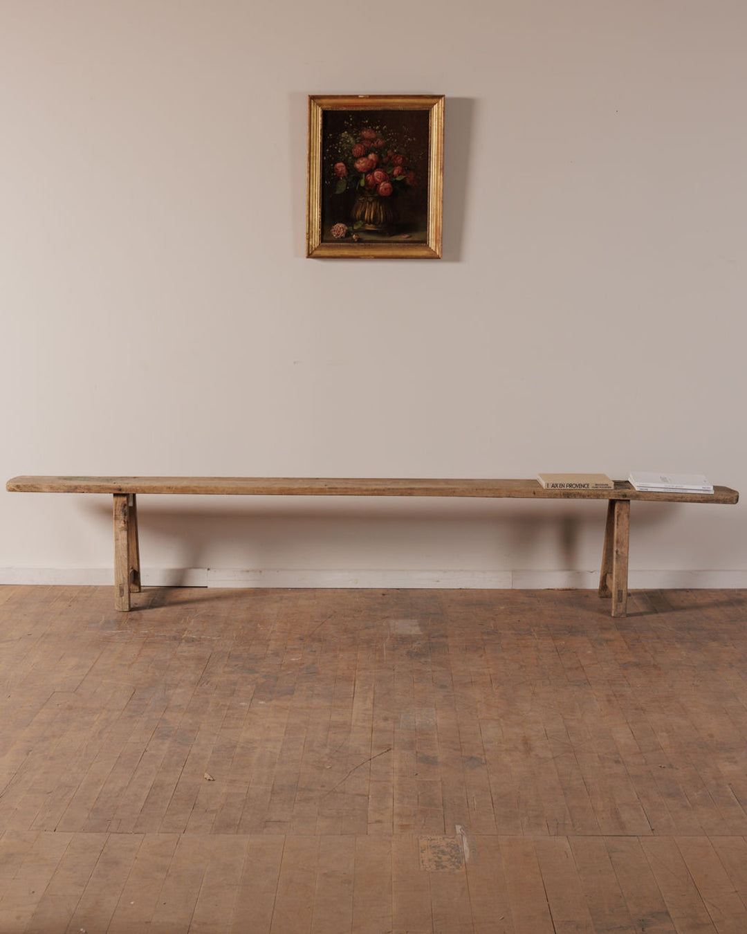 A long, light wooden bench with two books on its right end sits below a framed still life painting of red flowers on a pale wall.