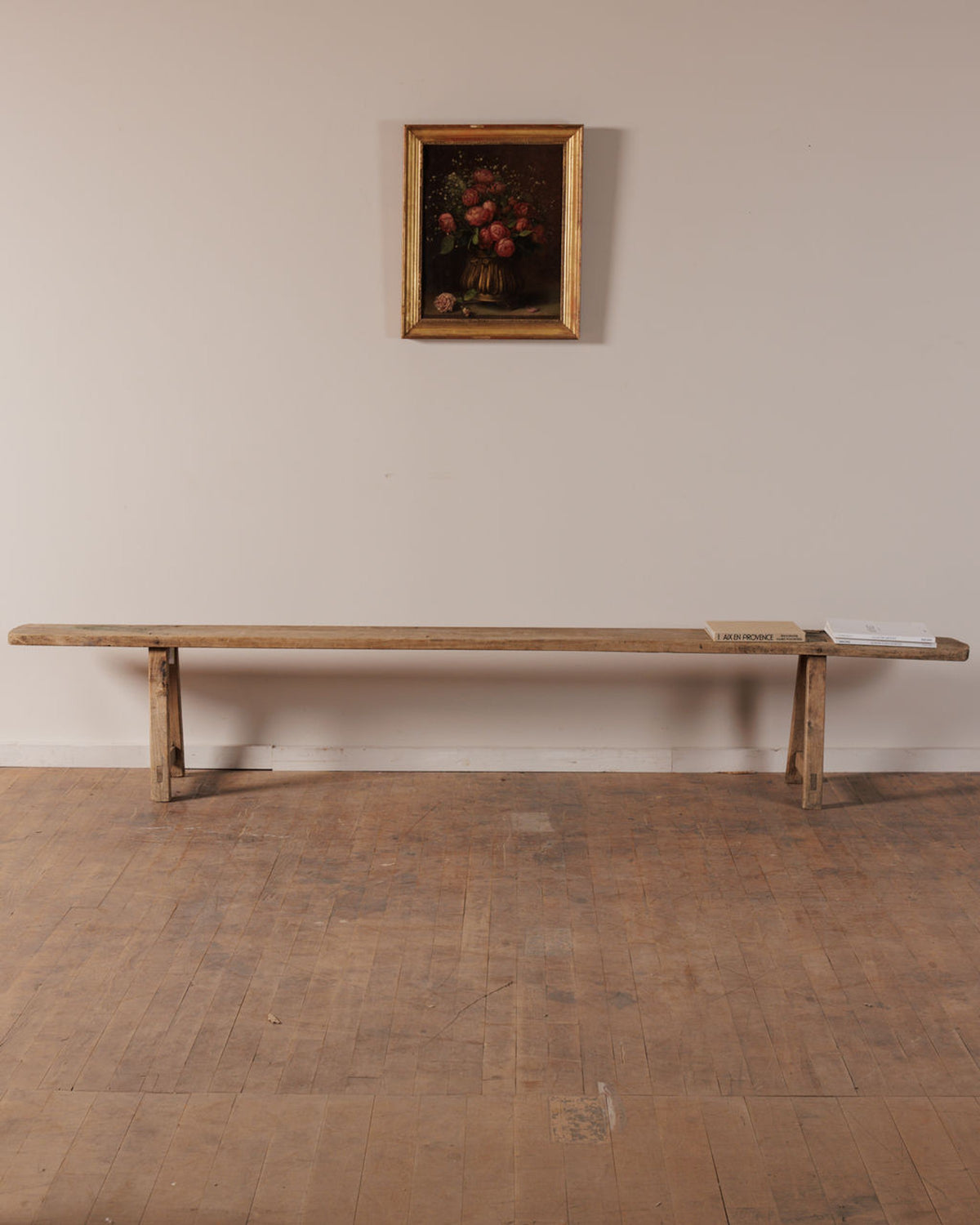 A long, light wooden bench with two books on its right end sits below a framed still life painting of red flowers on a pale wall.