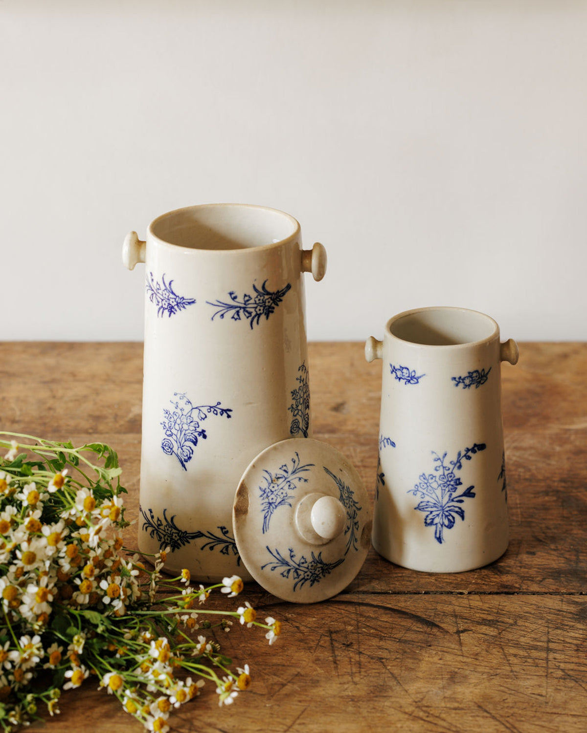 Two cream-colored ceramic jars with blue floral patterns, a matching lid, and a bunch of chamomile flowers on a wooden table.