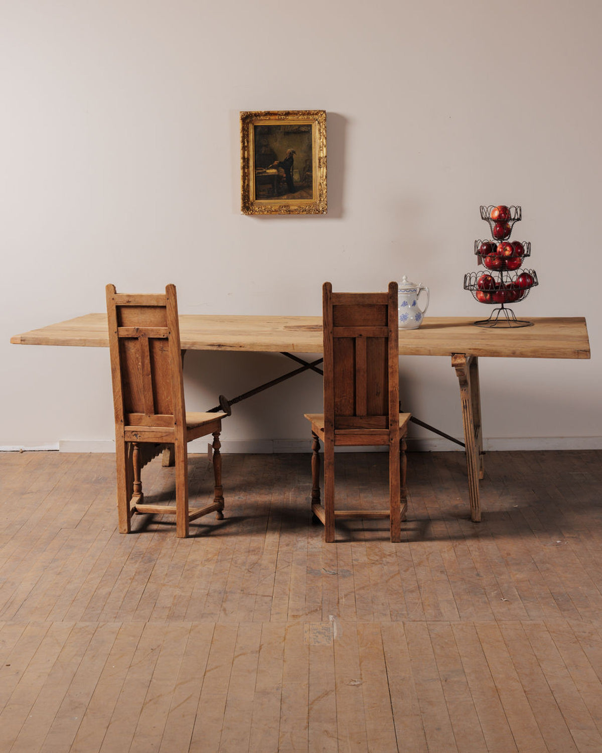 A long wooden dining table with two chairs, a framed painting, and a tiered apple basket.