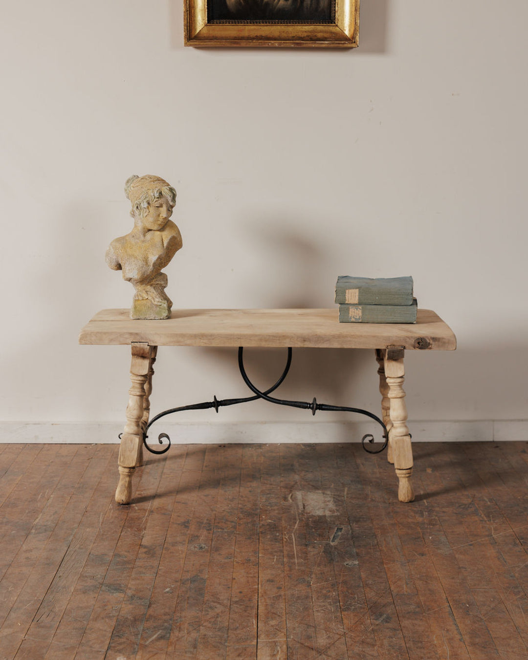 An antique light wood table with a wrought iron stretcher holds a bust and two books on a wood floor, beneath a framed painting.
