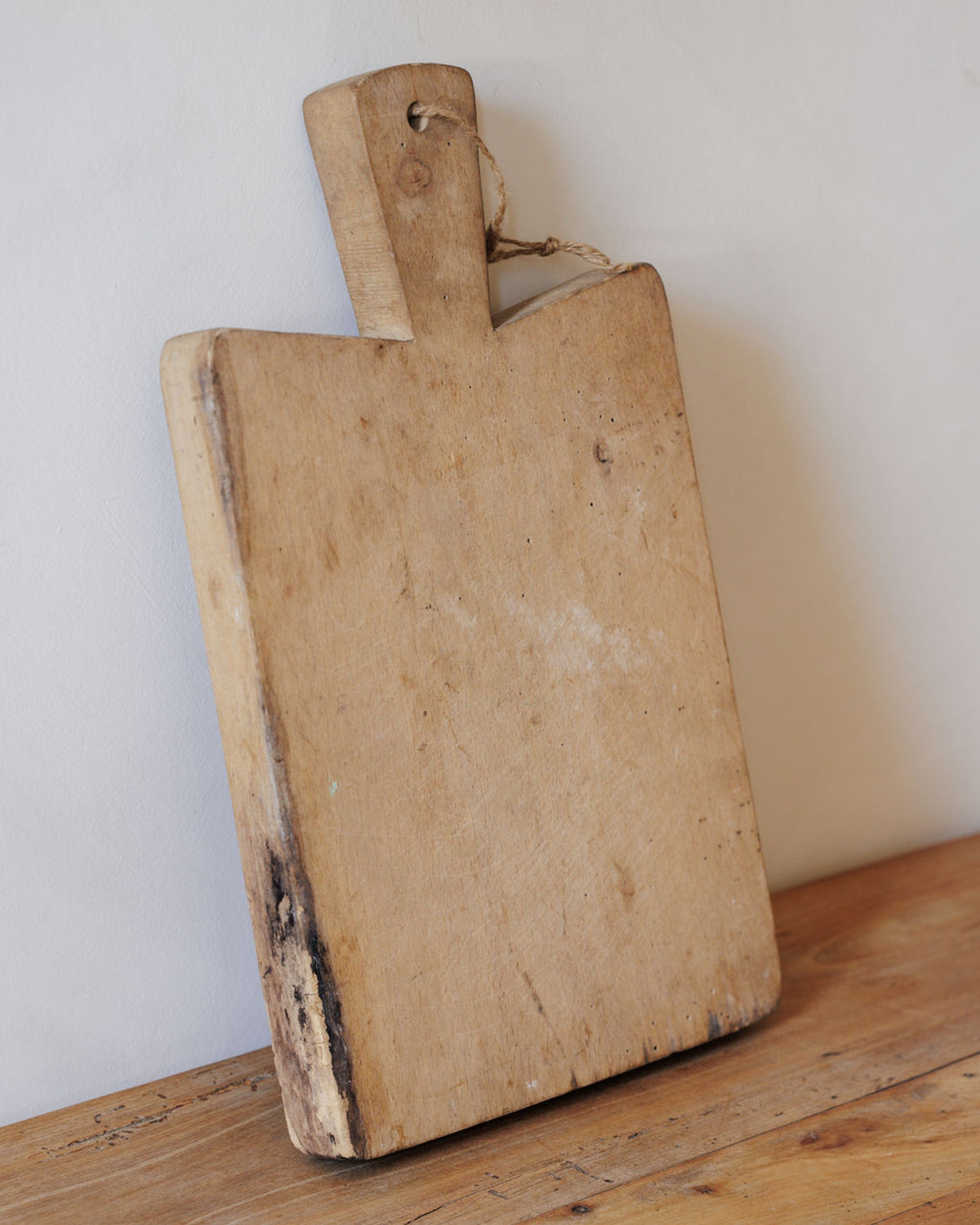 An old, worn wooden cutting board with a twine loop leans against a wall on a wooden surface.