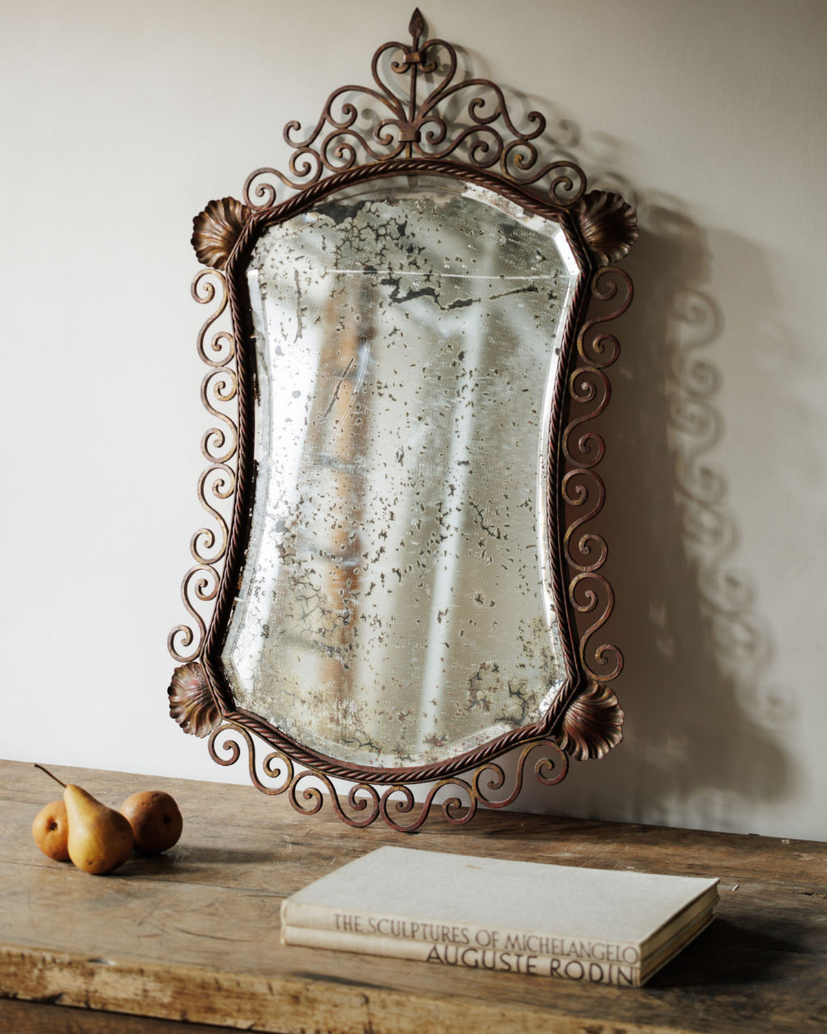 Ornate antique mirror with distressed glass and dark scrolled frame, on a rustic wooden table with pears and a book.